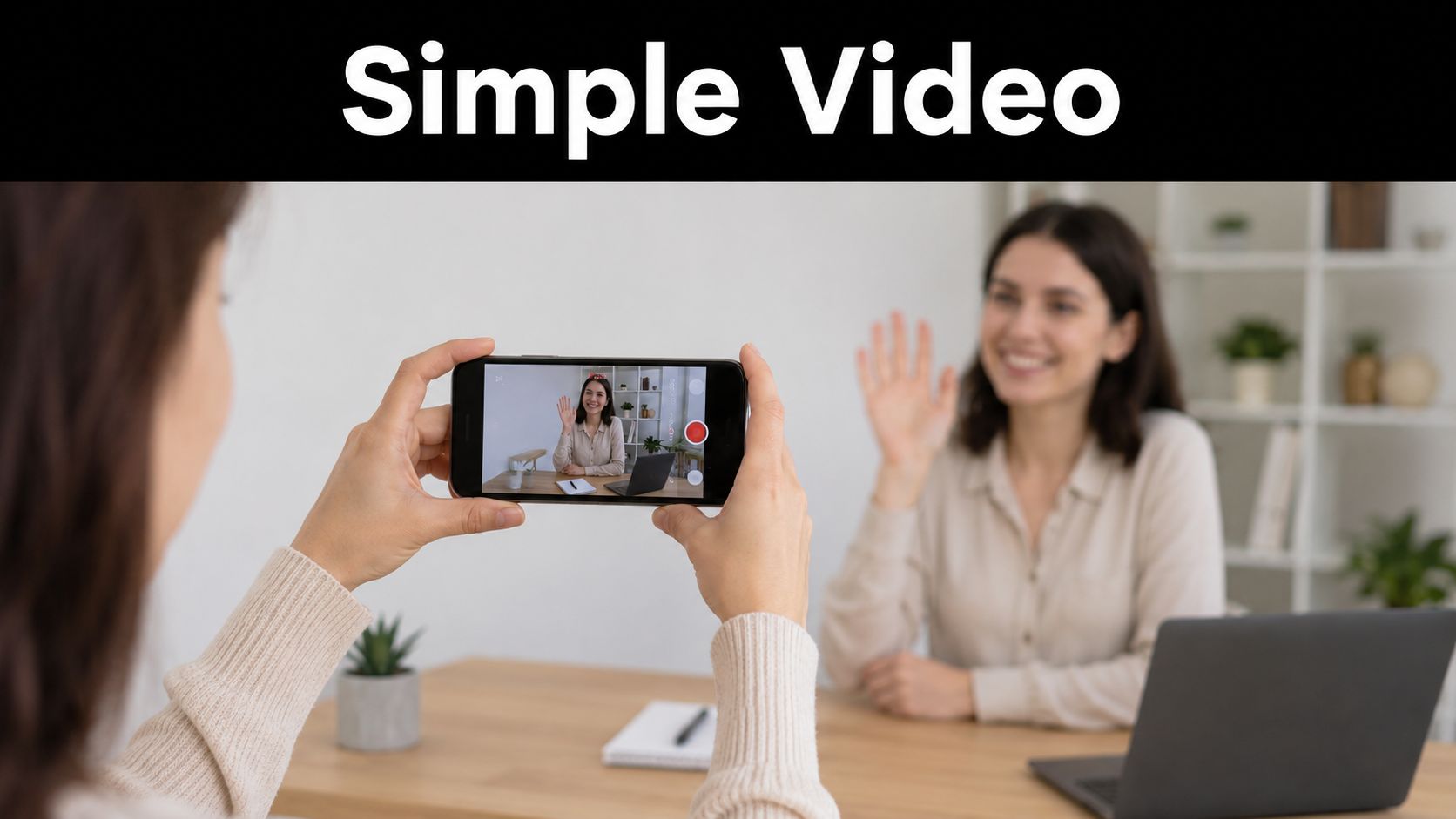 A person filming a woman waving at a laptop camera using a smartphone in an office.